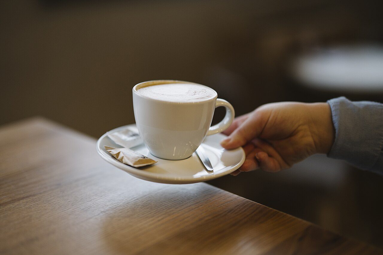 Secangkir kopi hangat di atas meja kayu yang dipegang oleh tangan seseorang, salah satu minuman berkafein yang perlu dibatasi penderita asam lambung